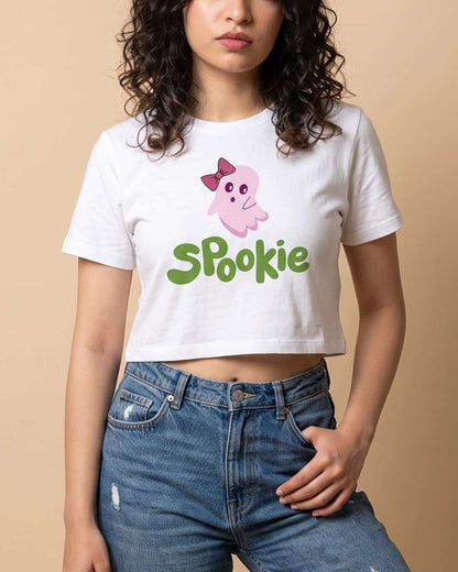 A woman wearing a white crop top t-shirt with a pink cartoon ghost and the word 'Spookie' in green, paired with blue jeans, against a solid beige background.